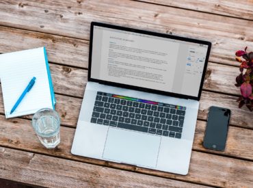 aufgeklappter Laptop steht auf einem Tisch, daneben ein Glas, Papier und ein Stift
